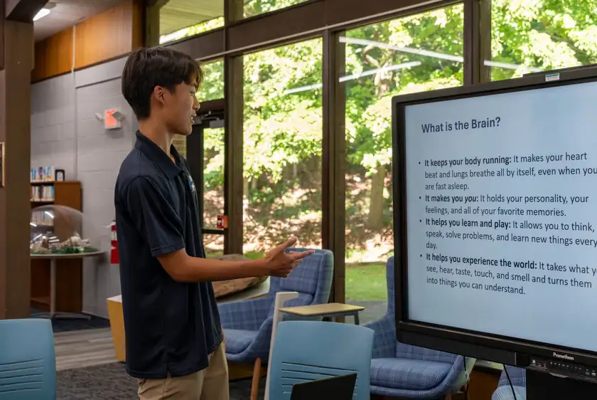 A presenter points to a large interactive display showing a slide titled &ldquo;What is the Brain?&rdquo; in a library room with tall windows