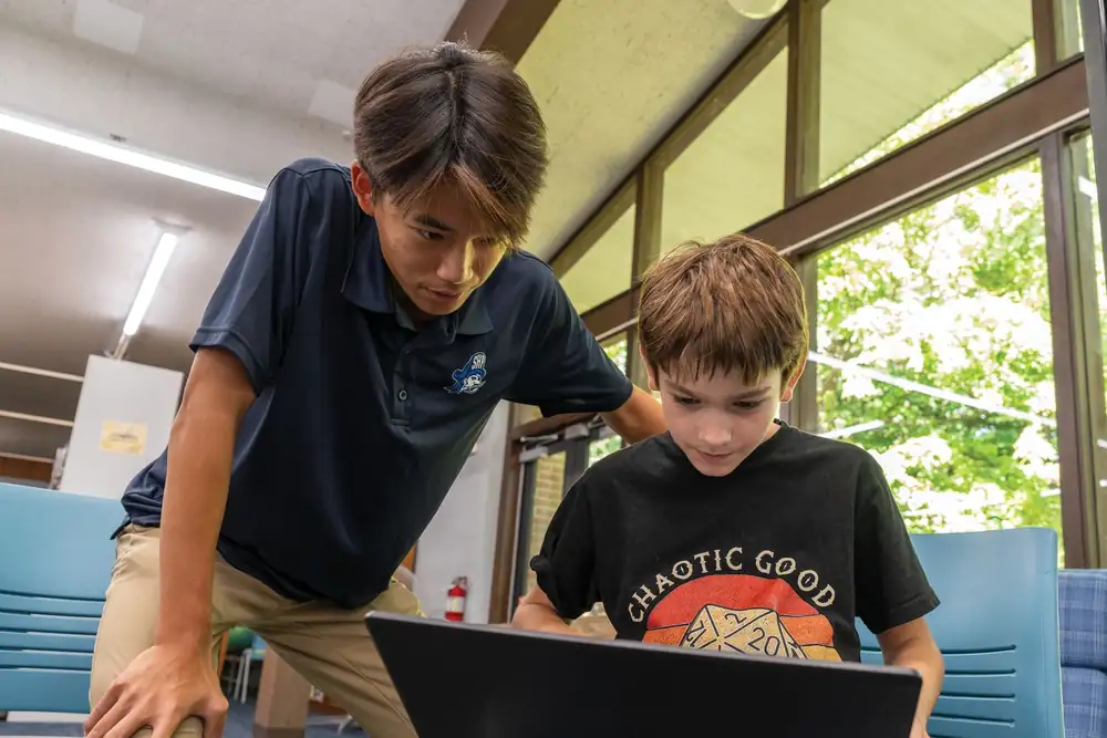 A presenter leans in beside a young student who is working on a laptop in a library